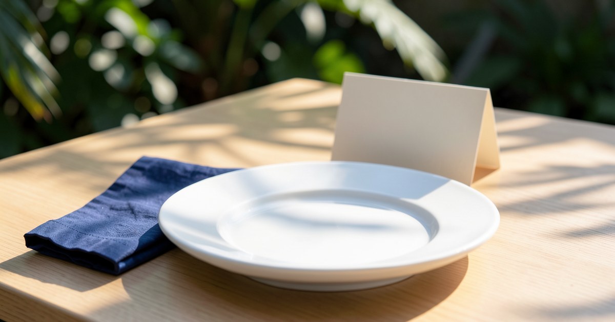White dinner plate, navy napkin and blank place card on a sunlit wooden table.